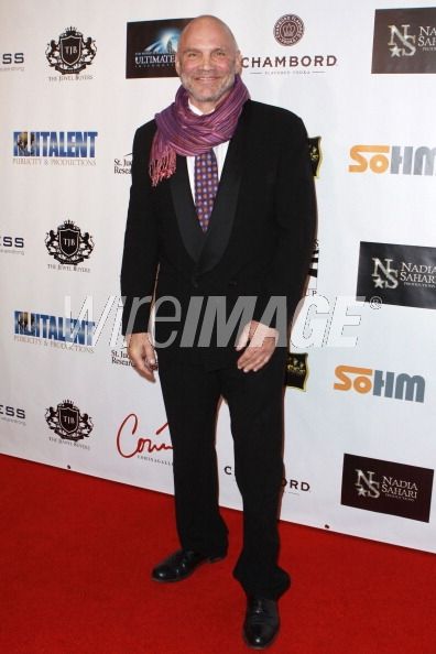 Man in a black suit with a purple scarf standing on a red carpet in front of a branded backdrop.