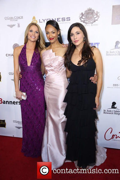 Three women in formal dresses standing on a red carpet with a branded backdrop.
