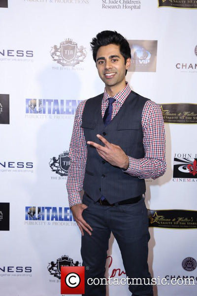 Smiling man in a dress shirt, tie, and waistcoat standing against a white event sponsor wall.