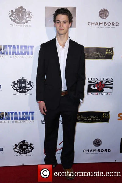 Man in a black suit standing on a red carpet with event logos in the background