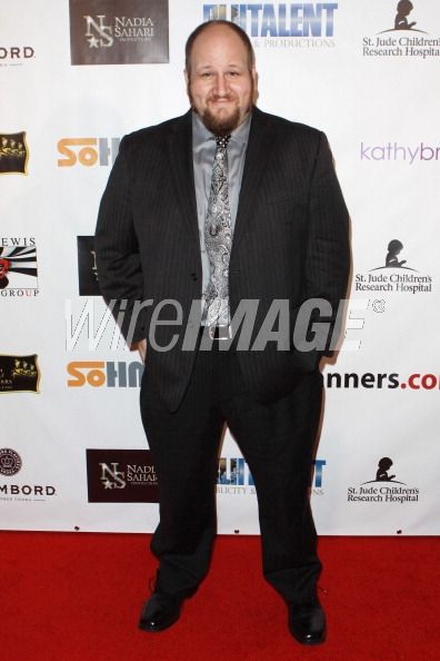 Man in a suit standing on a red carpet with event sponsor logos in the background