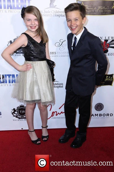 Two children on a red carpet event with a backdrop featuring logos and text.