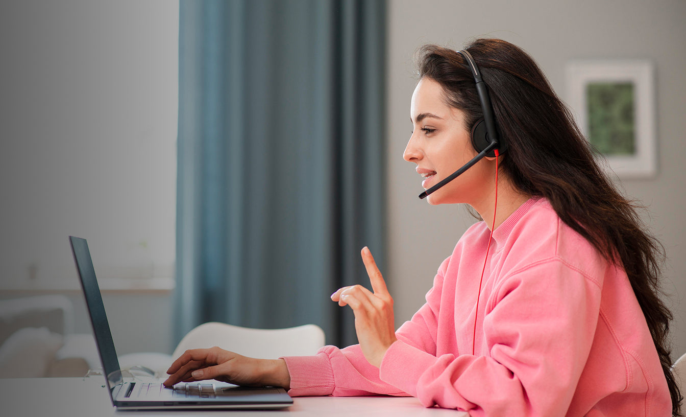 Woman in pink sweatshirt using a laptop with a headset, sitting in a room with a curtain and picture frame.